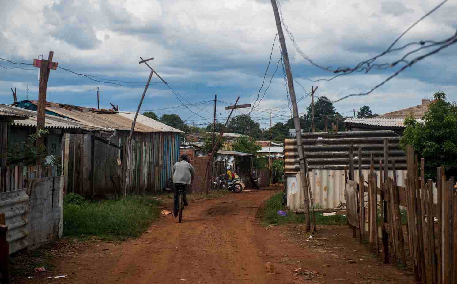Homem pedala em rua de terra entre casas de madeira e chapas metálicas